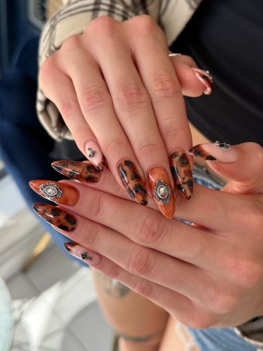 Long, stylish nails with a mix of orange and black designs, featuring rhinestones.