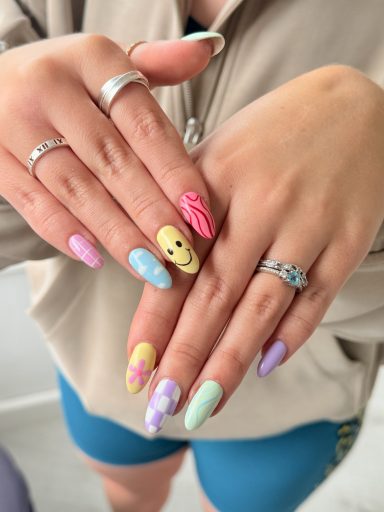 Colourful nails with designs, including a smiley face and floral patterns, displayed with rings.